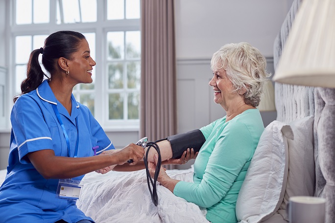 Senior Woman At Home In Bed Having Blood Pressure Taken By Female Home Healthcare Worker In Uniform